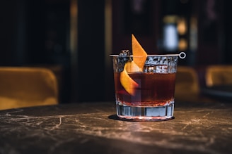 A cocktail served in a short glass with a garnish, placed on a textured stone or marble surface in a dimly lit ambiance. The drink has a rich, amber hue complemented by a twist of orange peel and a cocktail cherry, helping to create an elegant and sophisticated setting.