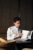 A person with long hair wearing a white shirt is sitting at a table, deeply focused on reading a magazine. The setting appears to be a stylish, possibly upscale indoor environment with subdued lighting, contributing to a cozy and intimate atmosphere. A glass of white wine is placed on the table, adding an element of sophistication.