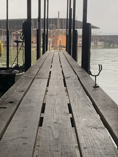 A long wooden dock stretches towards a body of water, with several vertical poles on either side. The background features buildings and structures that appear to be workshops or warehouses near the water's edge. The scene has an industrial and somewhat serene setting.