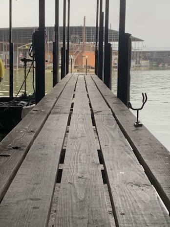 A long wooden dock stretches towards a body of water, with several vertical poles on either side. The background features buildings and structures that appear to be workshops or warehouses near the water's edge. The scene has an industrial and somewhat serene setting.