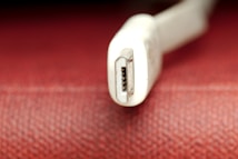 A close-up view of a white USB connector, possibly a Mini USB, set against a textured red background. The focus is on the metal connector, which has a slightly worn appearance.