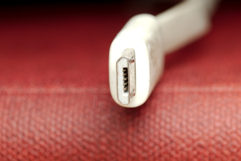 A close-up view of a white USB connector, possibly a Mini USB, set against a textured red background. The focus is on the metal connector, which has a slightly worn appearance.