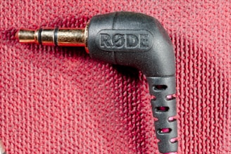A close-up image of a black audio jack connector labeled with the brand 'RODE' against a textured red fabric background. The connector is at a right angle, and the metallic plug is visible.