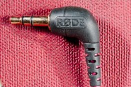 A close-up image of a black audio jack connector labeled with the brand 'RODE' against a textured red fabric background. The connector is at a right angle, and the metallic plug is visible.