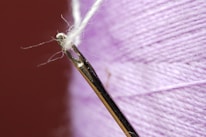 A close-up view of a threaded needle with a delicate white thread. The background features a spool of purple thread displaying its texture.