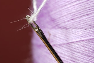 A close-up view of a threaded needle with a delicate white thread. The background features a spool of purple thread displaying its texture.