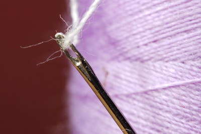 A close-up view of a threaded needle with a delicate white thread. The background features a spool of purple thread displaying its texture.