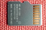 A close-up image of a microSD card lying on a textured red surface. The card has white printed text including alphanumeric codes and the words 'MADE IN PHILIPPINES'. On the right side, several gold-colored contact points are visible.