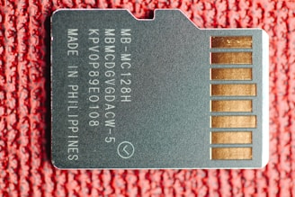 A close-up image of a microSD card lying on a textured red surface. The card has white printed text including alphanumeric codes and the words 'MADE IN PHILIPPINES'. On the right side, several gold-colored contact points are visible.