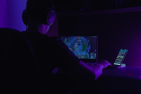 A person wearing headphones is sitting at a desk, focused on a computer screen displaying a video game. There is also a smartphone placed on a stand next to the screen, illuminated by a purple ambient light.