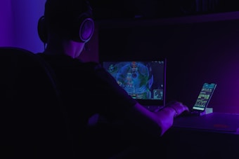 A person wearing headphones is sitting at a desk, focused on a computer screen displaying a video game. There is also a smartphone placed on a stand next to the screen, illuminated by a purple ambient light.