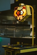 An arcade game machine is situated indoors, featuring a sign labeled '4P VS' with a retro design including characters reminiscent of classic video games. The cabinet is predominantly yellow with colored joysticks.