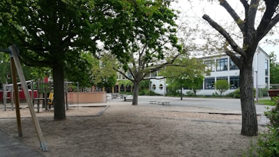 A playground with various equipment surrounded by large trees and a sandy area. In the background, there is a modern white building with large windows and visible greenery. The playground appears empty, and the setting suggests a peaceful environment.
