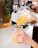 Hands are pouring a bright orange blended beverage from a blender into a glass jar. A whole pineapple and a sliced orange are visible on a kitchen counter in the background.