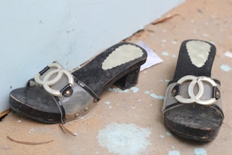 A pair of black heeled sandals with transparent straps and metallic double-loop emblems placed on a slightly worn and dirty ground surface. The background includes a light blue wall, with a piece of paper partially visible under one of the sandals.