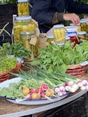 A market stall features a variety of fresh produce and preserves. There are jars filled with pickled vegetables like peppers, aligned on a table. Fresh greens, radishes, spring onions, and other leafy vegetables are neatly arranged on plates and baskets. A person is partially visible, appearing to be tending the stall.