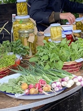 A market stall features a variety of fresh produce and preserves. There are jars filled with pickled vegetables like peppers, aligned on a table. Fresh greens, radishes, spring onions, and other leafy vegetables are neatly arranged on plates and baskets. A person is partially visible, appearing to be tending the stall.