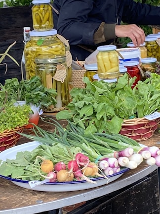A market stall features a variety of fresh produce and preserves. There are jars filled with pickled vegetables like peppers, aligned on a table. Fresh greens, radishes, spring onions, and other leafy vegetables are neatly arranged on plates and baskets. A person is partially visible, appearing to be tending the stall.