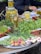 A market stall features a variety of fresh produce and preserves. There are jars filled with pickled vegetables like peppers, aligned on a table. Fresh greens, radishes, spring onions, and other leafy vegetables are neatly arranged on plates and baskets. A person is partially visible, appearing to be tending the stall.
