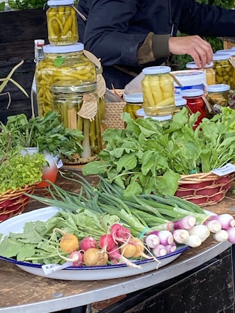 A market stall features a variety of fresh produce and preserves. There are jars filled with pickled vegetables like peppers, aligned on a table. Fresh greens, radishes, spring onions, and other leafy vegetables are neatly arranged on plates and baskets. A person is partially visible, appearing to be tending the stall.