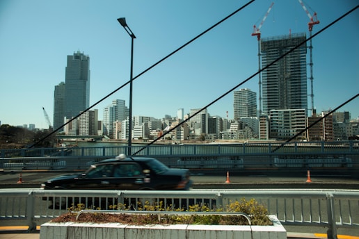 An urban cityscape featuring modern high-rise buildings and a construction site with cranes. A black taxi is driving across a bridge in the foreground. Several cables from the bridge are visible, stretching diagonally across the scene. The sky is clear, and there are some traffic cones along the road.