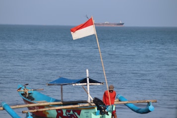 A small traditional fishing boat with bright turquoise and blue colors is anchored near the shore. A person in a red shirt is sitting on the boat, and an Indonesian flag is prominently displayed on a pole. In the distance, a large cargo ship is visible on the horizon.