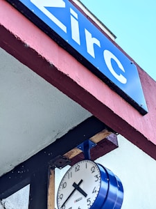 A blue sign with white text displaying the word 'Zirc' is mounted on a red and white overhang. Below the sign, a clock with black numbers and hands, encased in a blue frame, is attached to the structure.
