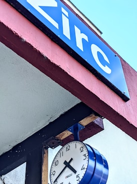 A blue sign with white text displaying the word 'Zirc' is mounted on a red and white overhang. Below the sign, a clock with black numbers and hands, encased in a blue frame, is attached to the structure.