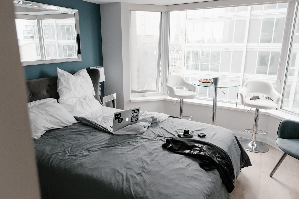 Urban studio apartment bedroom with dark grey bedding, teal accent wall, laptop on bed, and corner windows overlooking city buildings