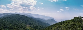 A panoramic view of lush green hills with dense forest covering them stretches out towards misty, distant mountains under a sky dotted with fluffy clouds. The horizon fades into a soft blue haze, giving a sense of depth and tranquility. On the right edge, part of a structure is visible, possibly a house or observation point.