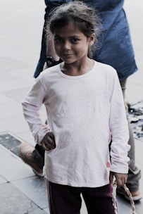 A young girl with a slight smile stands with one hand on her hip. Her long hair is loosely tied back, and she is wearing a white long-sleeve shirt with small embellishments paired with dark pants. She is standing on a tiled surface outdoors, with a person's legs visible behind her.