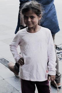 A young girl with a slight smile stands with one hand on her hip. Her long hair is loosely tied back, and she is wearing a white long-sleeve shirt with small embellishments paired with dark pants. She is standing on a tiled surface outdoors, with a person's legs visible behind her.