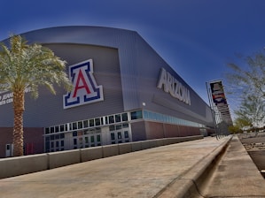 A large sports arena with the word 'Arizona' prominently displayed on the side. The building is modern with a large letter 'A' logo. There is a palm tree in the foreground, and banners are displayed on the right side along a walkway. The setting is sunny with blue skies.