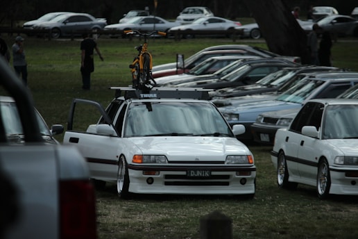 A parking lot with several vintage cars lined up, predominantly white in color. In the foreground, a prominently featured vehicle has a bicycle mounted on its roof. The scene is set in a park area with a grassy ground and some trees visible. A few people are walking in the background.