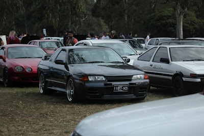A group of classic cars are parked closely together on a grassy area with people walking in the background. The central car is a black vintage model with a license plate 1187-H3, flanked by a red sports car on the left and a white coupe on the right. The scene takes place in a park-like setting with trees around.