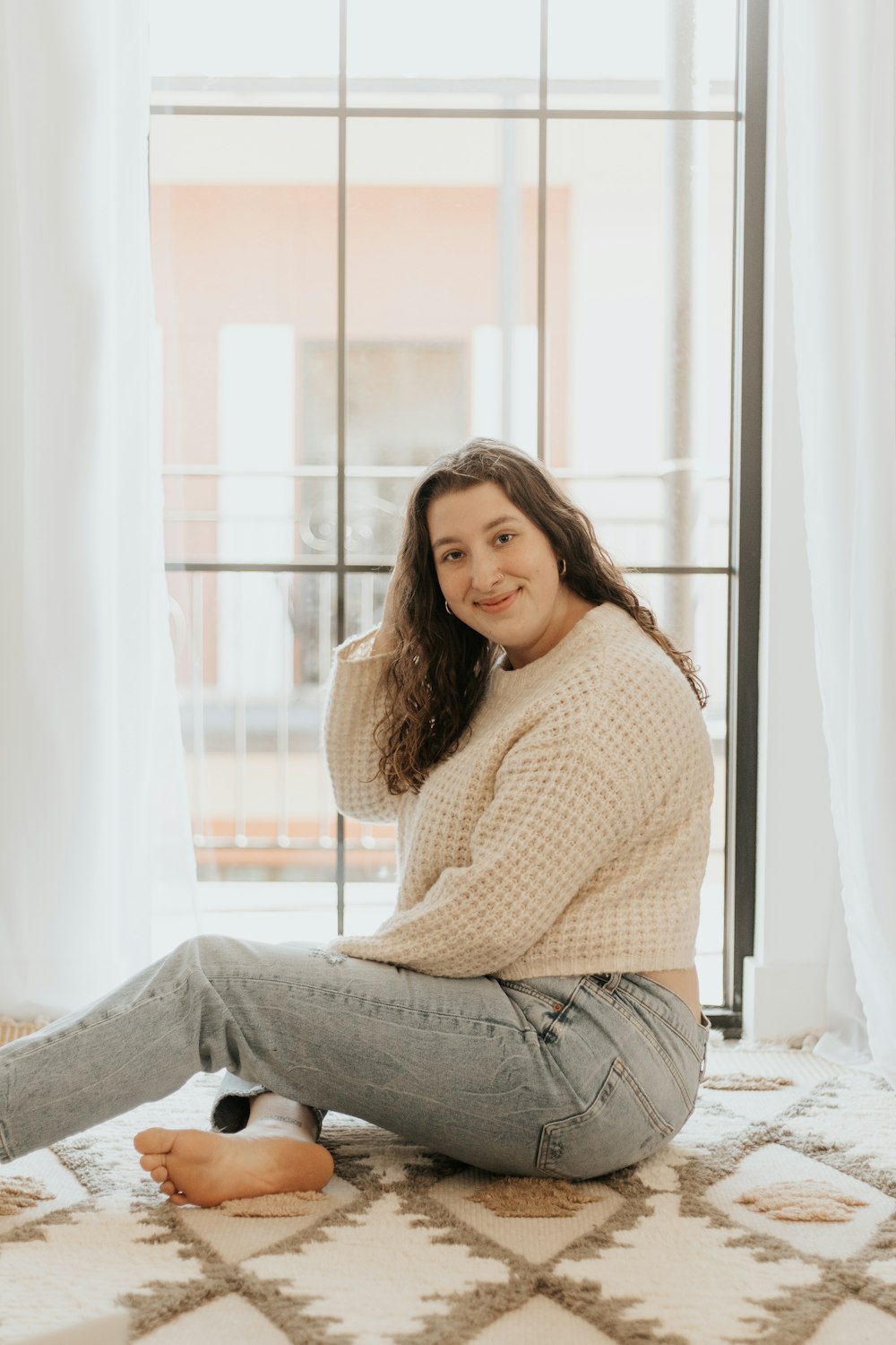 woman in brown sweater and gray denim jeans sitting on white window curtain