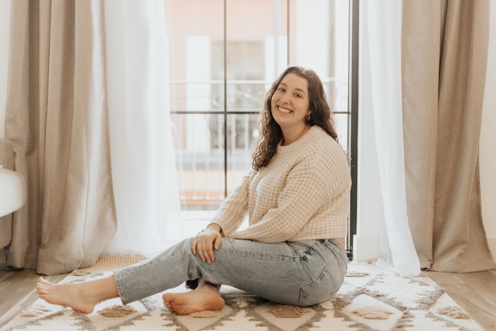 Frau in weißem Strickpullover und grauer Jeans sitzt auf weißem und braunem Blumenbett