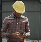 man in red blue and white plaid dress shirt wearing yellow hat holding black smartphone