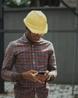 man in red blue and white plaid dress shirt wearing yellow hat holding black smartphone