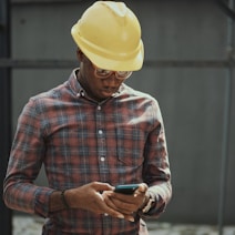man in red blue and white plaid dress shirt wearing yellow hat holding black smartphone