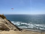 A paraglider soaring above a vibrant coastline with waves crashing below.
