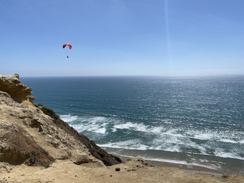 Paraglider soaring over a coastal mountain landscape under clear skies.
