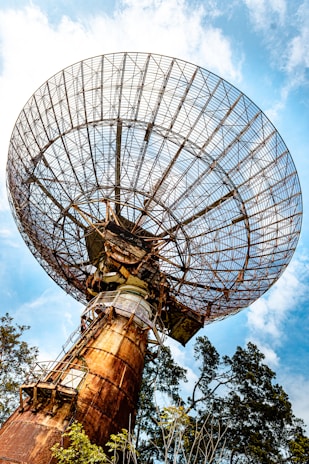 A large, rusted satellite dish extends upwards, dominating the scene against a backdrop of a clear blue sky with a few scattered clouds. The dish's intricate metal framework is prominently displayed, with trees partially visible at the base, emphasizing the juxtaposition of technology and nature.