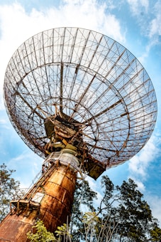 A large, rusted satellite dish extends upwards, dominating the scene against a backdrop of a clear blue sky with a few scattered clouds. The dish's intricate metal framework is prominently displayed, with trees partially visible at the base, emphasizing the juxtaposition of technology and nature.