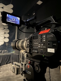 A professional video camera is set up on a tripod in a dimly lit studio or stage setting. The camera is equipped with various buttons, displays, and a large lens. In the background, there are cases and sound equipment, indicating a production environment.