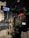 A professional video camera is set up on a tripod in a dimly lit studio or stage setting. The camera is equipped with various buttons, displays, and a large lens. In the background, there are cases and sound equipment, indicating a production environment.