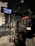 A professional video camera is set up on a tripod in a dimly lit studio or stage setting. The camera is equipped with various buttons, displays, and a large lens. In the background, there are cases and sound equipment, indicating a production environment.