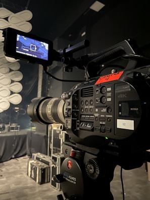 A professional video camera is set up on a tripod in a dimly lit studio or stage setting. The camera is equipped with various buttons, displays, and a large lens. In the background, there are cases and sound equipment, indicating a production environment.