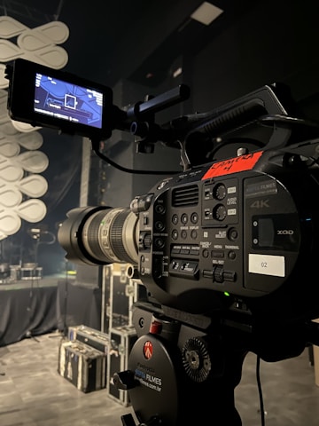 A professional video camera is set up on a tripod in a dimly lit studio or stage setting. The camera is equipped with various buttons, displays, and a large lens. In the background, there are cases and sound equipment, indicating a production environment.