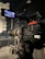 A professional video camera is set up on a tripod in a dimly lit studio or stage setting. The camera is equipped with various buttons, displays, and a large lens. In the background, there are cases and sound equipment, indicating a production environment.